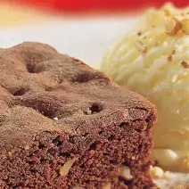 Fotografia em tons de marrom em uma bancada de madeira com uma foto aproximada de um prato polvilhado com cacau em pó, um brownie de chocolate e sorvete de creme.