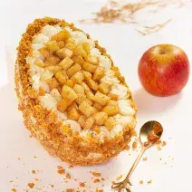 Foto da receita de Ovo Torta de Maçã, visto de cima, decorado com pedaços de maçã, massa de torta triturada, sobre uma bancada de mármore. Há uma maçã inteira e uma colher dourada na cena.
