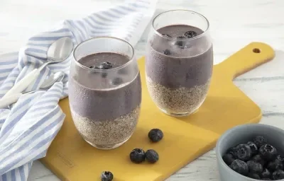 Receita de Pudim de Chia com Açaí e Mirtilos. Observa-se dois copos altos com uma parte de pudim de chia e outra parte de polpa de açaí, com mirtilos em cima. Os copos estão sobre uma tábua amarela e há um guardanapo de pano azul listrado sobre ela.