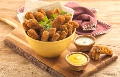 Fotografia em tons de amarelo em uma bancada de madeira clara, um paninho roxo, uma tábua de madeira com um potinho amarelo em cima e várias iscas empanadas de carne suína dentro dele. Ao lado, potinhos com mostarda e molho tártaro. Fotografia em tons de amarelo em uma bancada de madeira clara, um paninho roxo, uma tábua de madeira com um potinho amarelo em cima e várias iscas empanadas de carne suína dentro dele. Ao lado, potinhos com mostarda e molho tártaro.