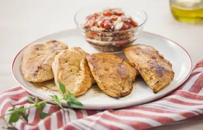 Fotografia em tons de vermelho em uma bancada de madeira clara com um pano branco com listras vermelhas, um prato branco raso com detalhes em vermelho e filés de frango dentro dele. Ao lado, um potinho de vidro com o molho vinagrete. Fotografia em tons de vermelho em uma bancada de madeira clara com um pano branco com listras vermelhas, um prato branco raso com detalhes em vermelho e filés de frango dentro dele. Ao lado, um potinho de vidro com o molho vinagrete.