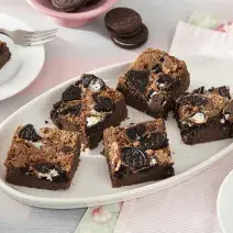 Foto da receita de Brownie de Leite Moça e Biscoito Negresco. Observa-se 7 fatias de brownie cortadas e dispostas em uma forma branca oval em um fundo rosa pastel.