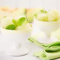 Fotografia em tons de verde em uma bancada de madeira de cor branca. Ao centro, uma taça contendo o sorvete e ao fundo há uma outra taça contendo sorvete. Ao lado, há um pano verde espalhado.