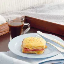 Fotografia em tons de amarela, marrom e rosa de uma bandeja de café da manhã em cima da cama, ao centro um prato branco redondo, dentro dele sanduíche, talheres e uma café preto. Abaixo do prato uma guardanapo azul claro xadrez para decorar.