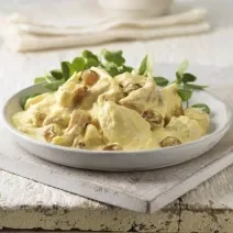 Fotografia em tons claros de uma salada de frango com molho ao curry e algumas folhas em um recipiente de cerâmica raso na cor branco. O prato está sobre uma peça de madeira em tom claro.
