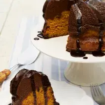 Fotografia em tons de marrom em uma bancada de madeira com um pano branco com listras azuis. Ao centro, um prato branco redondo raso com uma fatia do bolo de cenoura com chocolate. Ao fundo, um suporte branco para bolos com o bolo inteiro.