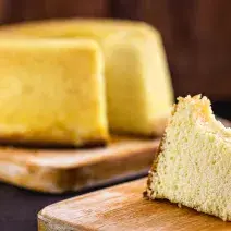 Fotografia em tons de marrom e amarelo de uma bancada azul escura vista de frente, duas tábuas de madeira uma com uma fatia de bolo e a outra com o bolo redondo sem uma fatia