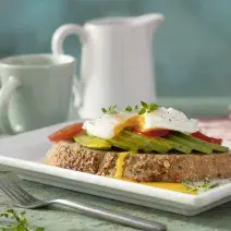 Fotografia em tons de vermelho em uma bancada de madeira verde clara, um paninho vermelho listrado, um prato quadrado branco com uma torrada e um ovo poché em cima com abacate. Ao fundo, tomates, xícara e jarra com café.