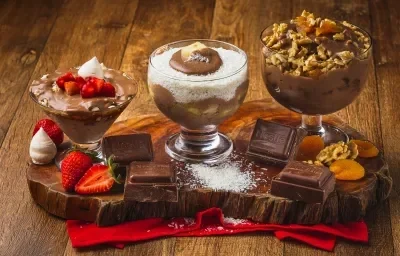 Foto da receita de Taças Cremosas, composta por três taças de vidro lado a lado sobre uma tábua de madeira com em um tecido vermelho. Cada uma está com um creme em tom marrom e alguns ingredientes compondo-as e a tábua está decorada. Foto da receita de Taças Cremosas, composta por três taças de vidro lado a lado sobre uma tábua de madeira com em um tecido vermelho. Cada uma está com um creme em tom marrom e alguns ingredientes compondo-as e a tábua está decorada.