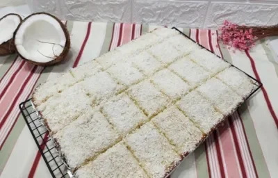 Foto da receita de Bolo Gelado com Coco. Observa-se uma grade preta com o bolo em cima cortado em retangulos e polvilhado de coco ralado seco. Foto da receita de Bolo Gelado com Coco. Observa-se uma grade preta com o bolo em cima cortado em retangulos e polvilhado de coco ralado seco.