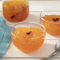Fotografia em tons de laranja em uma bancada de madeira branca, um pano azul, três potinhos de vidro com o doce de mamão ralado e em pedaços pequenos com cravos. Ao lado, duas colheres.