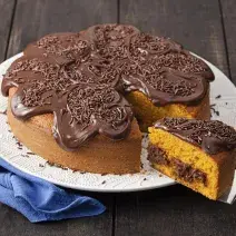 Fotografia em tons de marrom, azul e branco de uma bancada de madeira com um paninho azul e um prato redondo branco, sobre ele um bolo de cenoura com recheio e cobertura de chocolate .