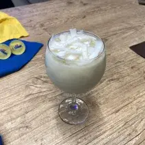 Fotografia em tons de amarelo e azul em uma bancada de madeira, com paninhos amarelo e azul e uma taça de vidro ao centro com a bebida de coco e Galak dentro, decorado com lascas de coco fresco.