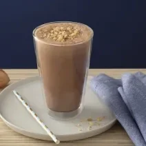 Foto em tons de marrom da receita de smoothie de café com nozes servida em um copo alto sobre uma base de porcelana cinza com um canudo listrado ao lado. Como decoração há duas nozes e um paninho azul