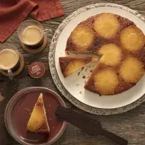 Foto de cima de um bolo invertido com rodelas de abacaxi, uma fatia está servida em um prato de sobremesa ao lado e duas xícaras transparentes com café preto e uma cápsula de Dolce Gusto Café Matinal.