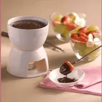 Fotografia de uma panela de fondue de cerâmica branca e aquecida com uma vela abaixo. Ao lado frutas frescas como morango, uva e banana em dois recipientes de vidro e um garfo próprio para fondue com uma morango coberto de chocolate.