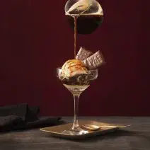 Fotografia em tons de preto de uma bancada de madeira com um prato quadrado, sobre ele uma taça com o affogato e uma jarra de café adicionando café á taça. Ao lado um paninho preto.