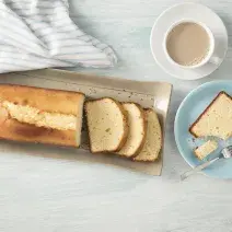 Fotografia em tons de azul em uma bancada de madeira clara com um prato redondo raso azul com uma fatia de bolo de Leite Moça. Ao lado, um recipiente retangular bege com o bolo inteiro.
