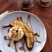 Fotografia vista de cima de uma banana cortada com meio com casca assada juntamente com uma bola de sorvete de creme. Por cima uma calda de caramelo desenhando todo prato, que é fundo e azul, em tom claro. O prato está sobre uma mesa marrom.