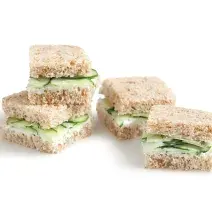 Fotografia em tons de verde em um fundo branco com quatro mini sanduichinhos no pão integral com recheio de pepino.