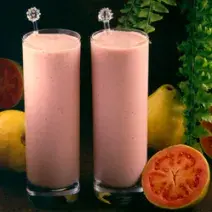 Fotografia em tons de rosa em uma bancada de madeira escura, dois copos de vidro alto com a bebida feita com suco de goiaba e Leite Moça. Ao lado, goiabas e uma goiaba aberta ao meio.