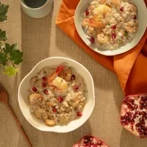 Foto da receita de risoto de camarão com brie e romã servida em um bowl de cerâmica branca sobre um paninho laranja e outro sobre uma mesa decorada com uma toalha de linho