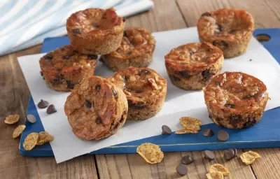 Fotografia em tons de azul em uma bancada de madeira com uma tábua azul, com um folha de papel manteiga e vários muffins de banana com gotas de chocolate em cima dela. Nesfit Cereais Matinais e algumas gotas de chocolate espalhadas pela bancada.