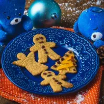 Fotografia em tons de laranja e azul em uma bancada de madeira, um paninho laranja, um prato azul com os biscoitinhos natalinos em cima dele. Ao lado, decoração e enfeites de natal.