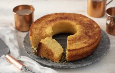 Fotografia em tons de dourado em uma bancada com uma toalha branca com detalhes cinzas, um prato redondo grande de pedra cinza e o bolo de queijo com coco em cima dele e uma fatia cortada. Ao lado, uma espátula com cabo cobre e ao fundo, várias xícaras.