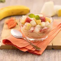 Foto da receita de salada de frutas servida em uma taça de vidro decorada com uma folha de hortelã, sobre um pano laranja com uma colher prateada ao lado. Ao fundo, há morangos, uma banana e fatia de melão