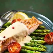 Fotografia de postas de salmão assado com um molho de iogurte e gengibre, por cima de alguns aspargos, limão siciliano do lado e tomates cereja assados. O salmão está em um prato azul raso, sobre um fundo verde escuro.