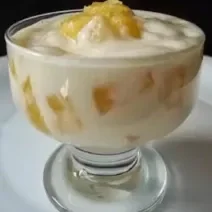 Foto da receita de mousse de abacaxi servida em uma taça de vidro com pedaços de abacaxi à mostra sobre um prato de porcelana branco