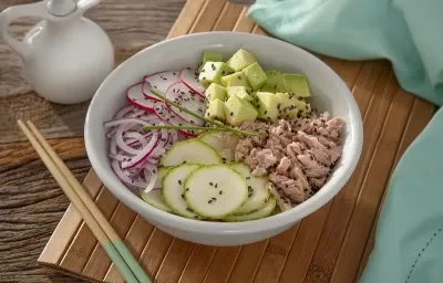 Foto em tons coloridos da receita de poke funcional de atum servida em um bowl de porcelana branco sobre uma esteira de madeira com dois hashis ao lado. Ao fundo há um pano azul claro