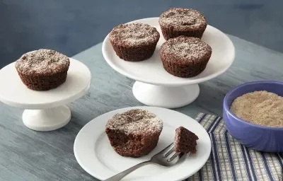 Foto em tons de cinza da receita de bolo de chocolate de farinha láctea aveia servida em diversas porções, três em uma base de porcelana branca e uma com uma garfada servida em um prato de mesmo material. Tudo isso sobre uma mesa cinza