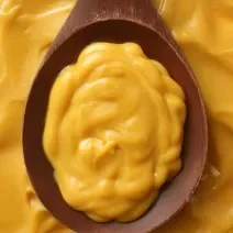 Fotografia vista de cima de um molho de mostarda em uma colher de madeira escura, apoiada sobre mais molho de mostarda.