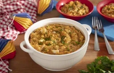 Foto da receita de Mungunzá de festa junina. Observa-se uma tigela grande branca com a receita dentro e, atrás, dois pratos fundos vermelhos cheios. Decoração de festa junina com tecidos e balões de papel decoram a foto do lado esquerdo