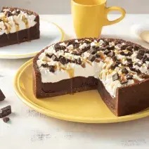 Fotografia em tons de amarelo em uma bancada branca com um prato grande amarelo e a torta de chocolate com caramelo em cima cortada ao meio. Ao lado, um prato branco com uma fatia da torta de chocolate, uma xícara amarela e pedaços de chocolates.