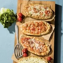 Fotografia vista de cima de quatro pães com linguiça e tomate, e outro com queijo, apoiados sobre papel manteiga, que estão sobre uma tábua de madeira. ao lado, tomates cereja, uma espátula pequena e um maço pequeno de alface.