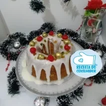 Fotografia em tons de vermelho em uma bancada de madeira de cor branca. Ao centro, o bolo contendo bolinhas coloridas em cima. Ao fundo, alguns enfeites natalinos na parede.