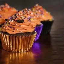 Foto de cupcakes cobertos por um creme laranja, decorados com confeitos coloridos em embalagens brilhantes nos tons de roxo e laranja.