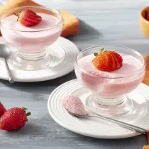 Imagem aproximada da receita de Mousse de Morango Zero Lactose, em tom rosa claro, servida em potes de vidro, decoradas com morangos fatiados. A receita está sobre pratos brancos em uma bancada com um tecido laranja e mais morangos inteiros decorando