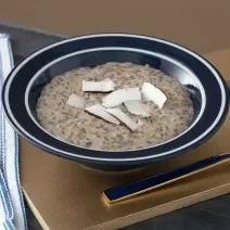 Foto em tons de marrom da receita de mingau de sementes e coco servida em um prato fundo preto com uma colher prateada ao lado sobre uma tábua de madeira lisa com um paninho azul ao lado