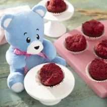 Foto da receita de Muffin de Beterraba. Observa-se uma tábua rosa com 6 muffins e, ao lado esquerdo, um pratinho branco com outro muffin rosa. O ursinho Bo está atrás desse pratinho branco.