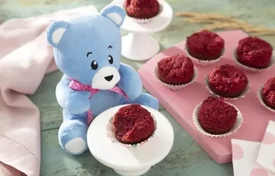 Foto da receita de Muffin de Beterraba. Observa-se uma tábua rosa com 6 muffins e, ao lado esquerdo, um pratinho branco com outro muffin rosa. O ursinho Bo está atrás desse pratinho branco.