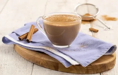 Foto da Receita de Cappuccino Cremoso. Observa-se uma xícara de vidro transparente sobre um guardanapo azul e uma tábua de madeira. Ao fundo, uma peneira com canela em pó.