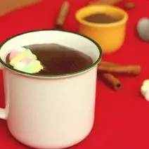 Fotografia em tons de branco e vermelho, ao centro uma xícara branca com chocolate quente e marshmallows, sobre toalha vermelha. Atrás um potinho com achocolatado e uma colher, com paus de canela e marshmallows espalhados ao redor.