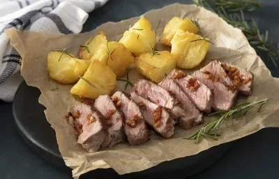 Foto em tons escuros da receita de bife ancho com batatas servidaem tiras de bife sobre um papel marrom em cima de uma pedra toda preta. Ao fundo há um pano branco quadriculado e ramos de alecrim