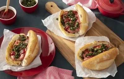 Foto em tons de vermelho da receita de choripan tradicional servida em três porções, duas sobre uma tábua de madeira e uma sobre um prato vermelho. Ao fundo há dois bowls de porcelana vermelhos além de uma panelinha e um paninho vermelhos