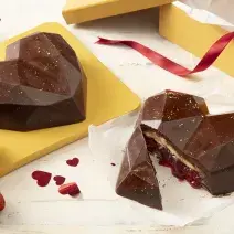 Fotografia em tons de marrom, amarelo e vermelho de uma bancada com dois corações de chocolate, um deles partido ao meio mostrando o recheio branco e de geleia de morango. Ao redor morangos e corações decorando.
