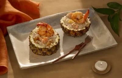Foto em tons de marrom da receita de risoto de camarão com abacaxi servida em duas porções em fatias de abacaxi sobre uma base de cerãmica cinza Foto em tons de marrom da receita de risoto de camarão com abacaxi servida em duas porções em fatias de abacaxi sobre uma base de cerãmica cinza